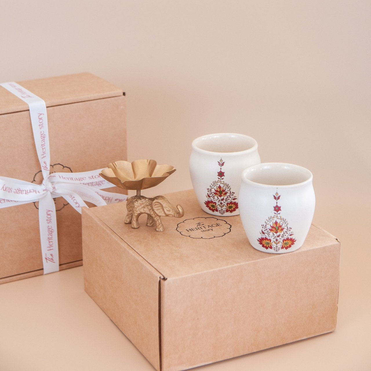 Two ceramic kulhads with floral designs and an elephant tealight on a cardboard gift box against a beige background
