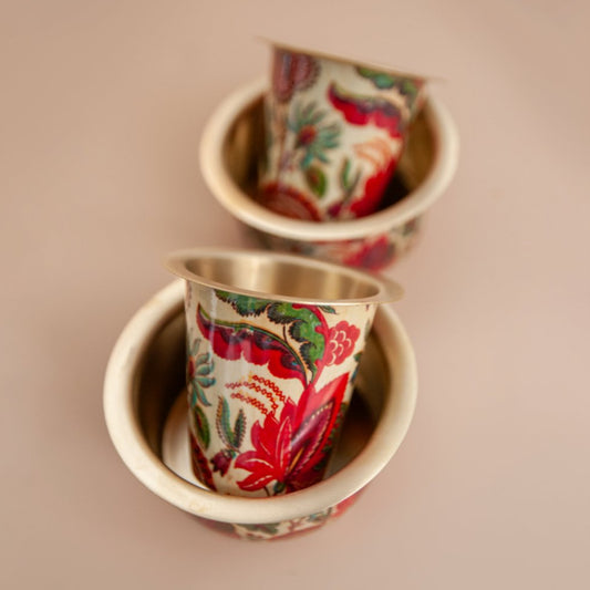 Two brass enameled south indian filter coffee set with floral patterns on a beige background