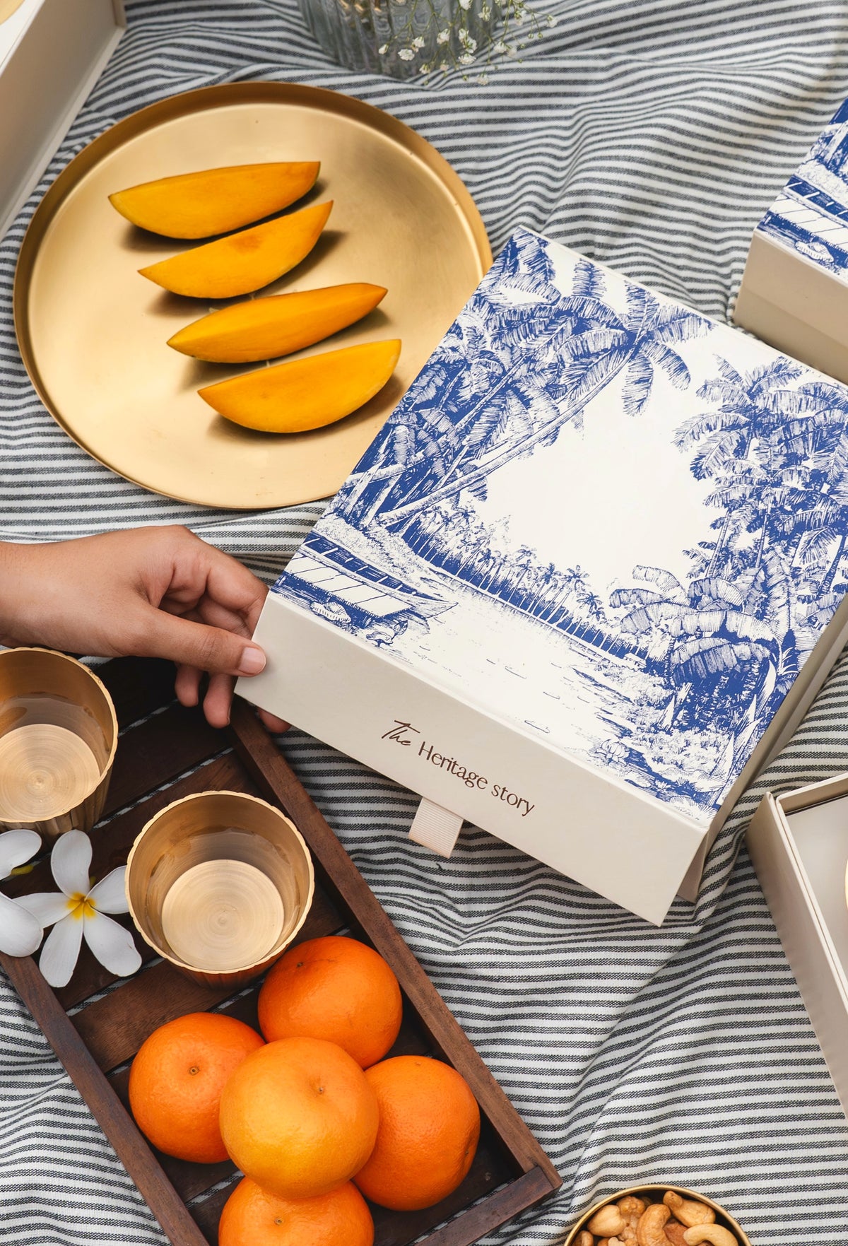 A hand holds a white gift box with blue scenic illustrations and the text 'Heritage Story' on a striped fabric surface. The scene includes sliced yellow fruit on a gold tray, whole oranges in a wooden tray, small metallic cups, and other gift packaging in a styled flat-lay arrangement.