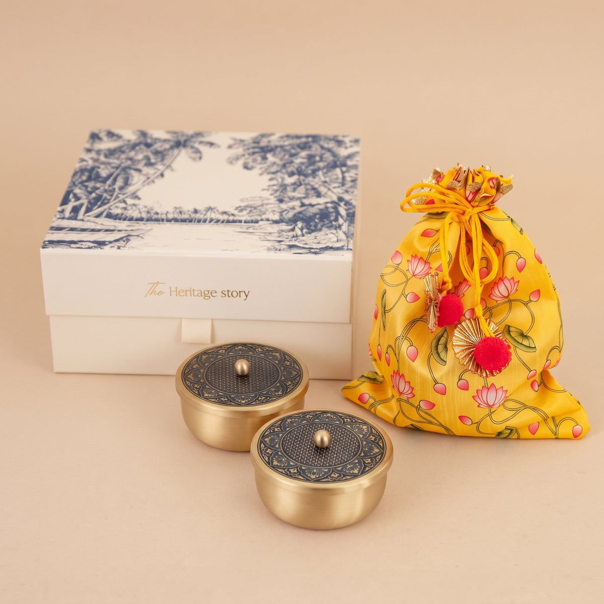 A gift set featuring two small round gold-toned metal containers with dark patterned lids, a yellow floral drawstring pouch with pink flowers and red pom-poms, and a white presentation box with blue scenic artwork and the text 'The Heritage story'.
