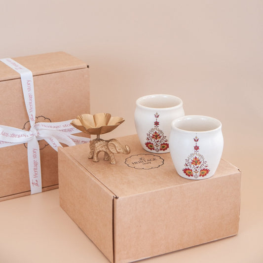 Two ceramic kulhads with floral designs and an elephant tealight on a cardboard gift box against a beige background