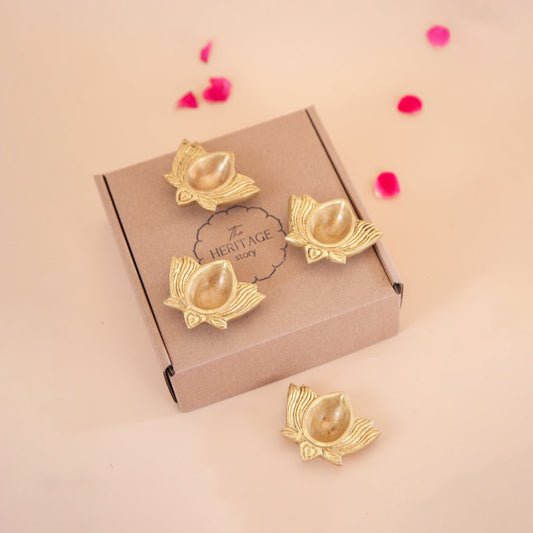 Brass lotus-shaped diyas (oil lamp) on a 'The Heritage Story' branded box with pink flower petals on a beige background.