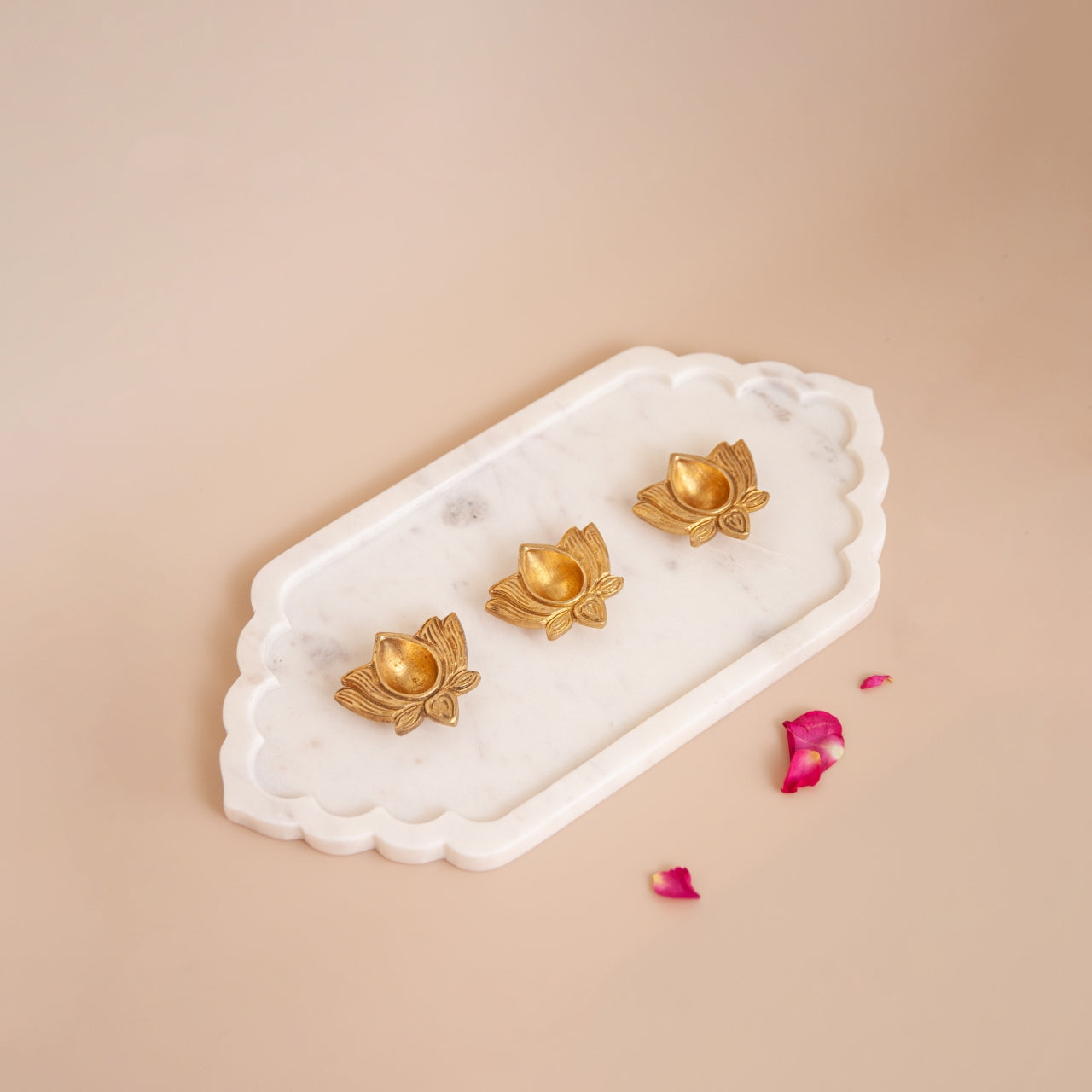 Gold lotus-shaped brass diyas on a decorative marble tray with pink petals on a beige background