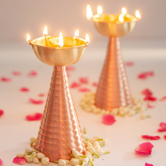 Two copper brass diya with lit candles on a light surface with scattered flowers.